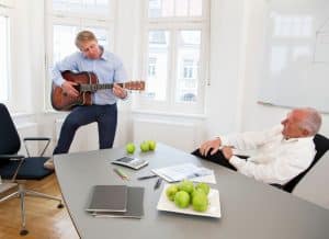 Mann spielt Gitarre in Büro und ein Kollege hört zu.