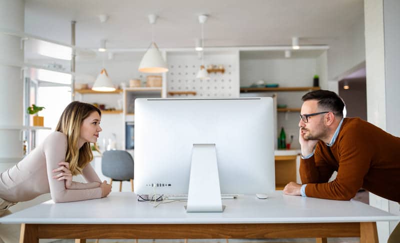 Zwei Mitarbeiter mit gleichem Job schauen sich auf einem Schreibtisch lehnend an und sprechen über Jobsharing