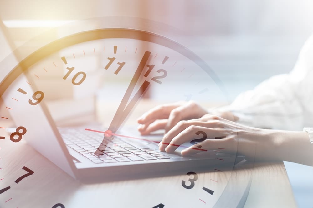 Eine transparente Uhr mit Zeiger und im Hintergrund ein Laptop. Arbeitszeit wird erfasst.